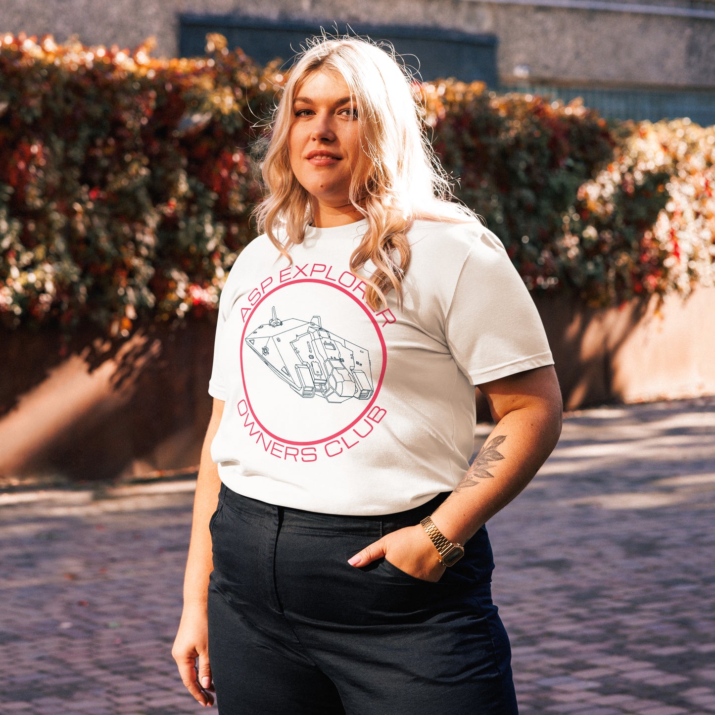 three quarters view of plus size woman wearing white Asp Explorer Owners club t-shirt in midnight blue and pink, with wireframe image of Asp explorer and text round a circle. Backdrop is outdoors, with a wall and vegetation out of focus.